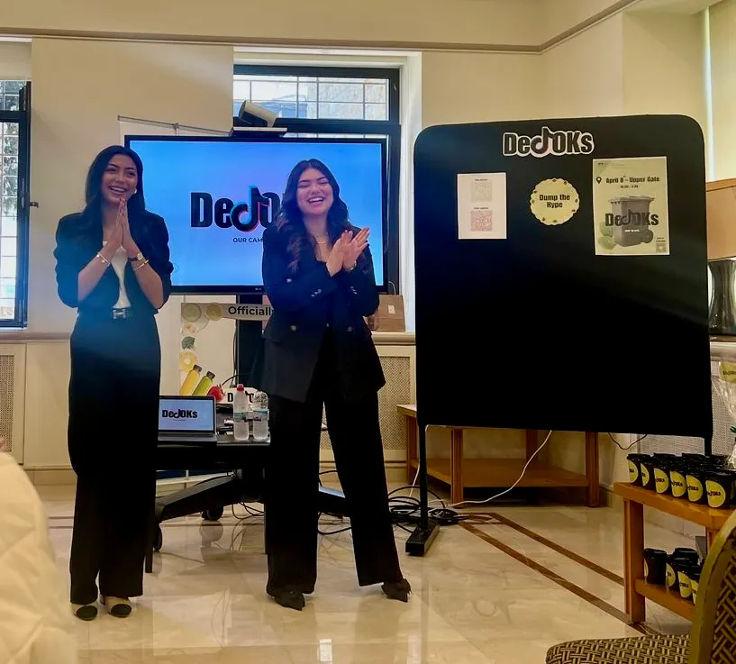 Joudi and Lea presenting DeTOKs in front of the campaign screen and the booth board.